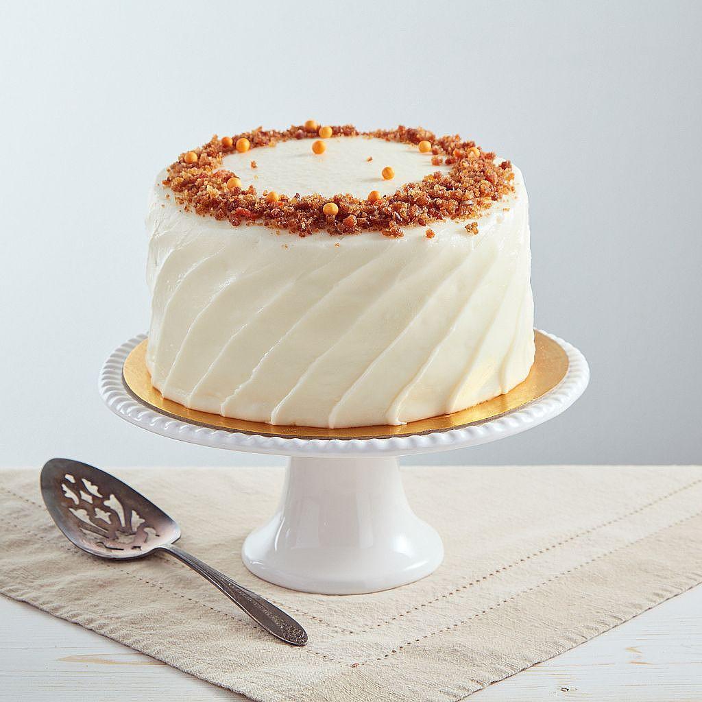 Six-inch carrot cake with swirled cream cheese frosting, topped with crunchy pecans and orange sprinkles on a white stand.