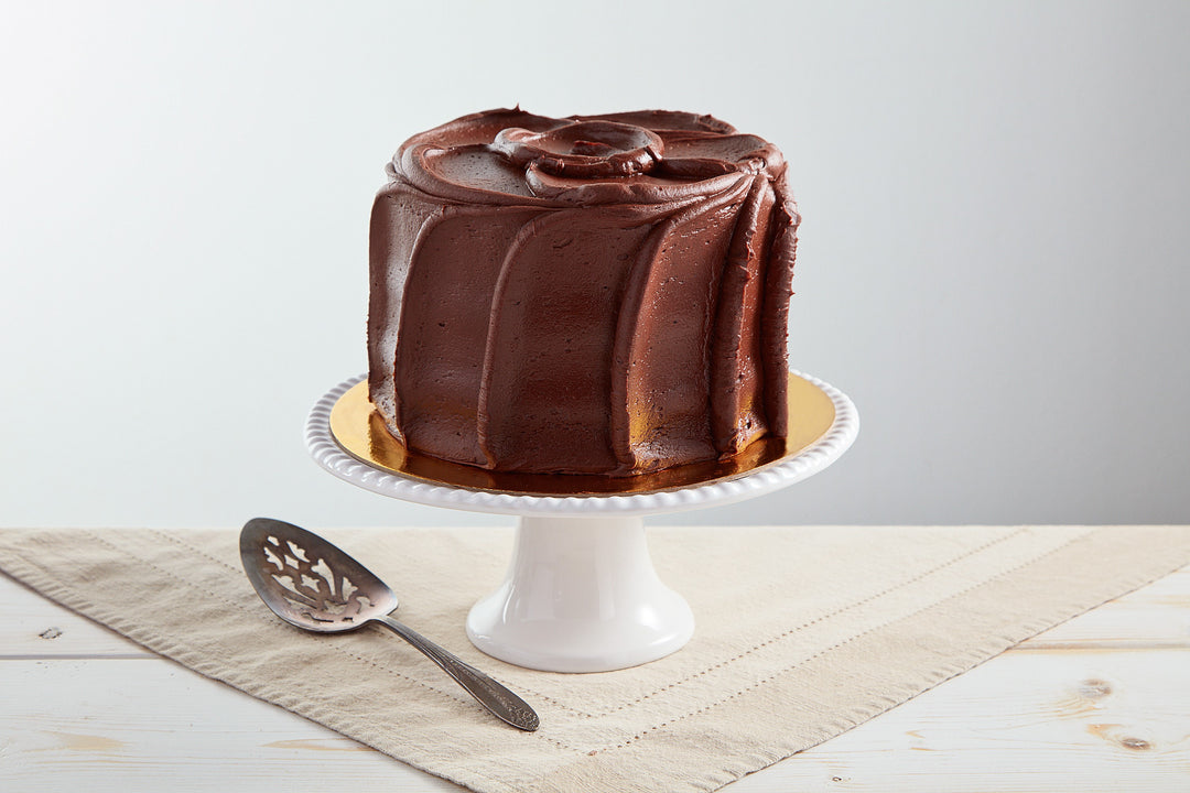 6-inch golden cake with rich chocolate buttercream frosting on a white pedestal stand with a silver cake server.