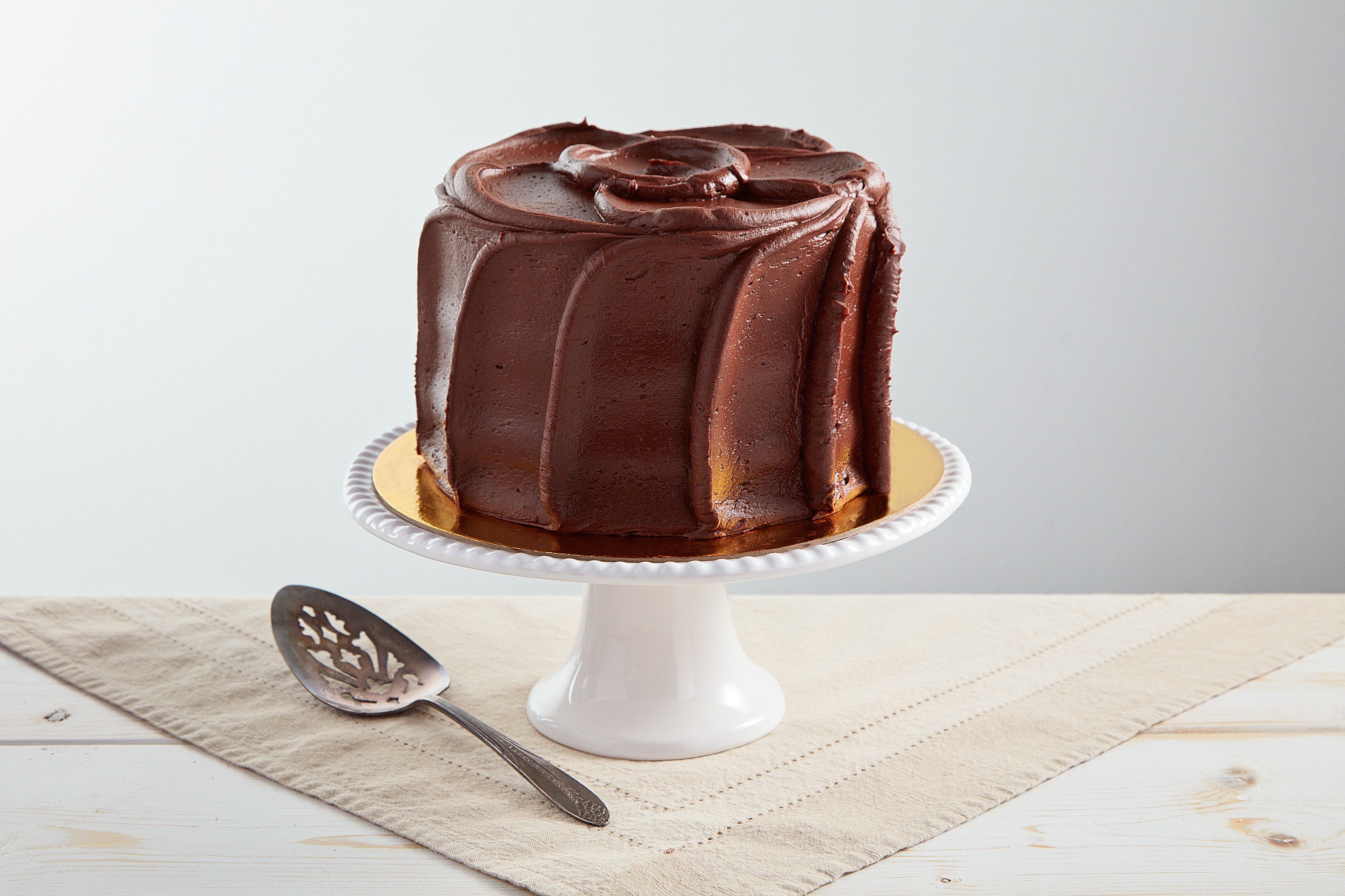 6-inch golden cake with rich chocolate buttercream frosting on a white pedestal stand with a silver cake server.