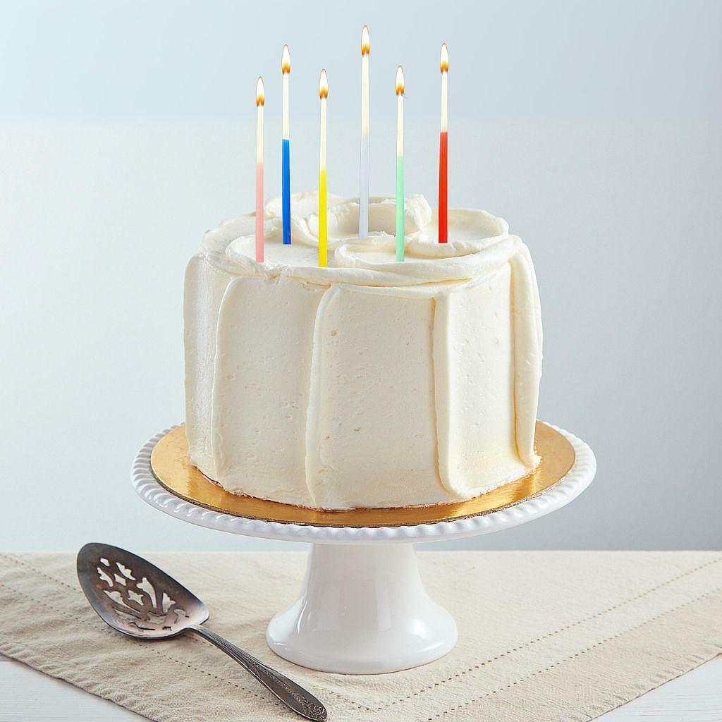 Six-inch French vanilla cake with smooth white buttercream frosting, topped with six lit colorful birthday candles on a white stand.