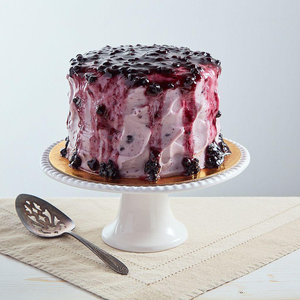 6-inch lemon blueberry cake with purple cream cheese frosting and a wild blueberry topping on a white cake stand.