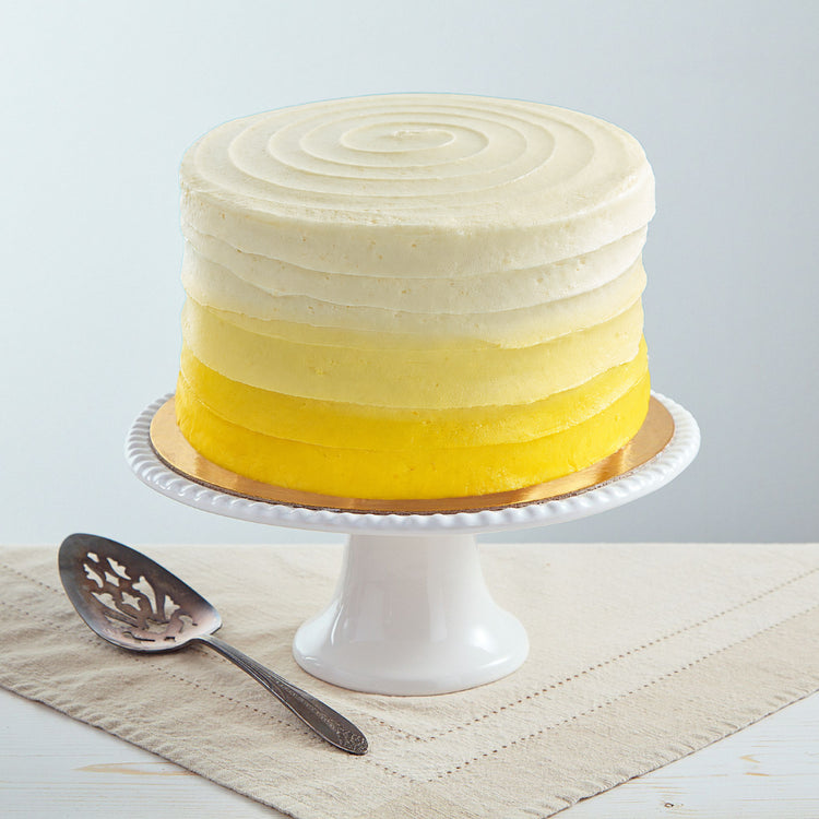 6-inch lemon cake with yellow ombre buttercream frosting on a white pedestal stand with a silver cake server.