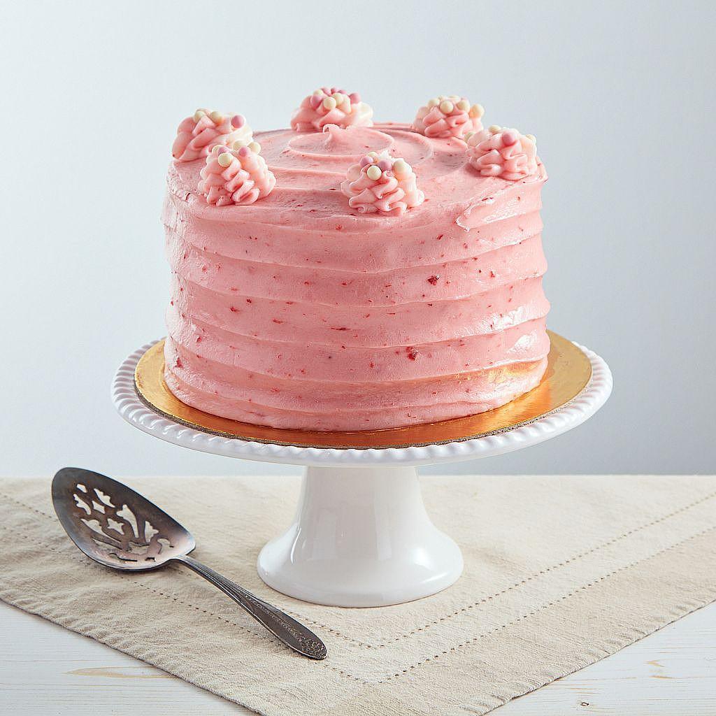 6-inch strawberry cake with pink cream cheese frosting, topped with piped swirls and sprinkles on a white pedestal stand.