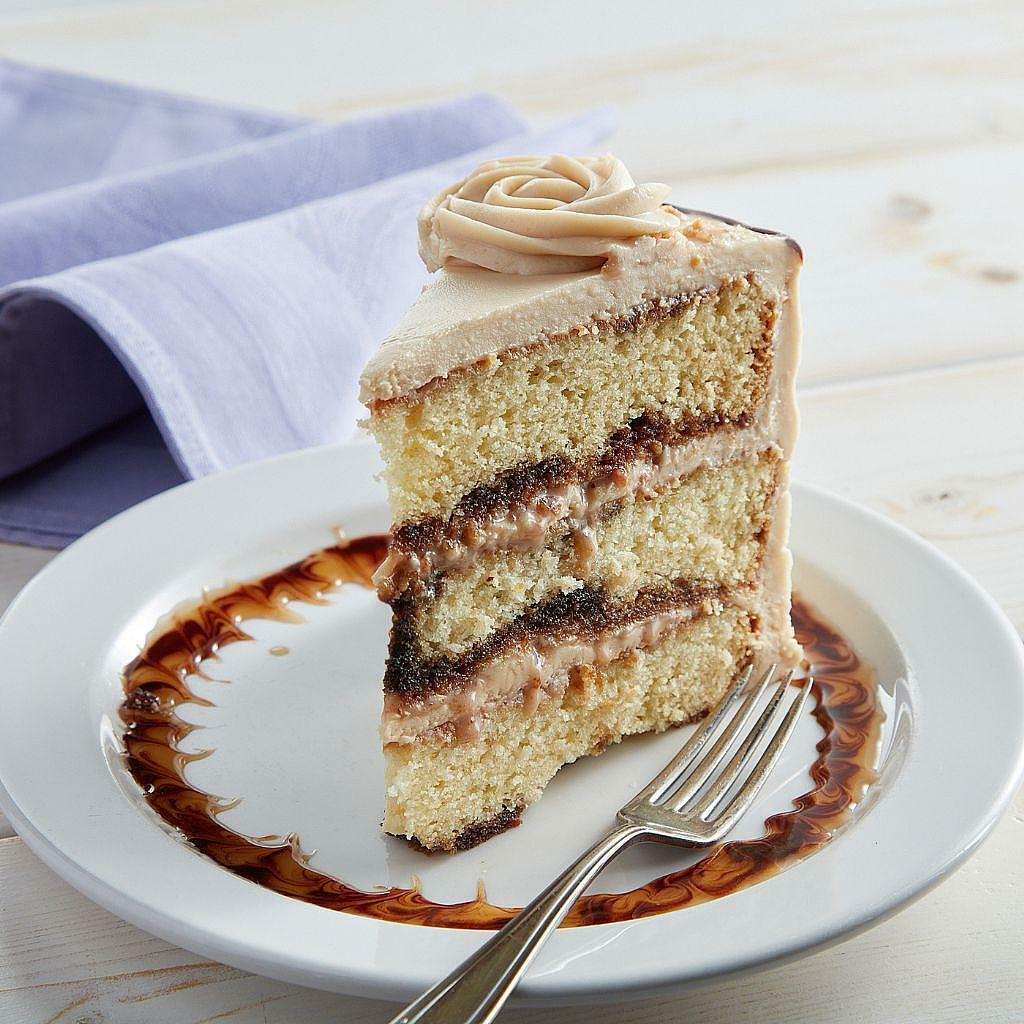 Slice of yellow layer cake with coffee frosting, toffee bits, and a decorative swirl on a white plate with a silver fork.
