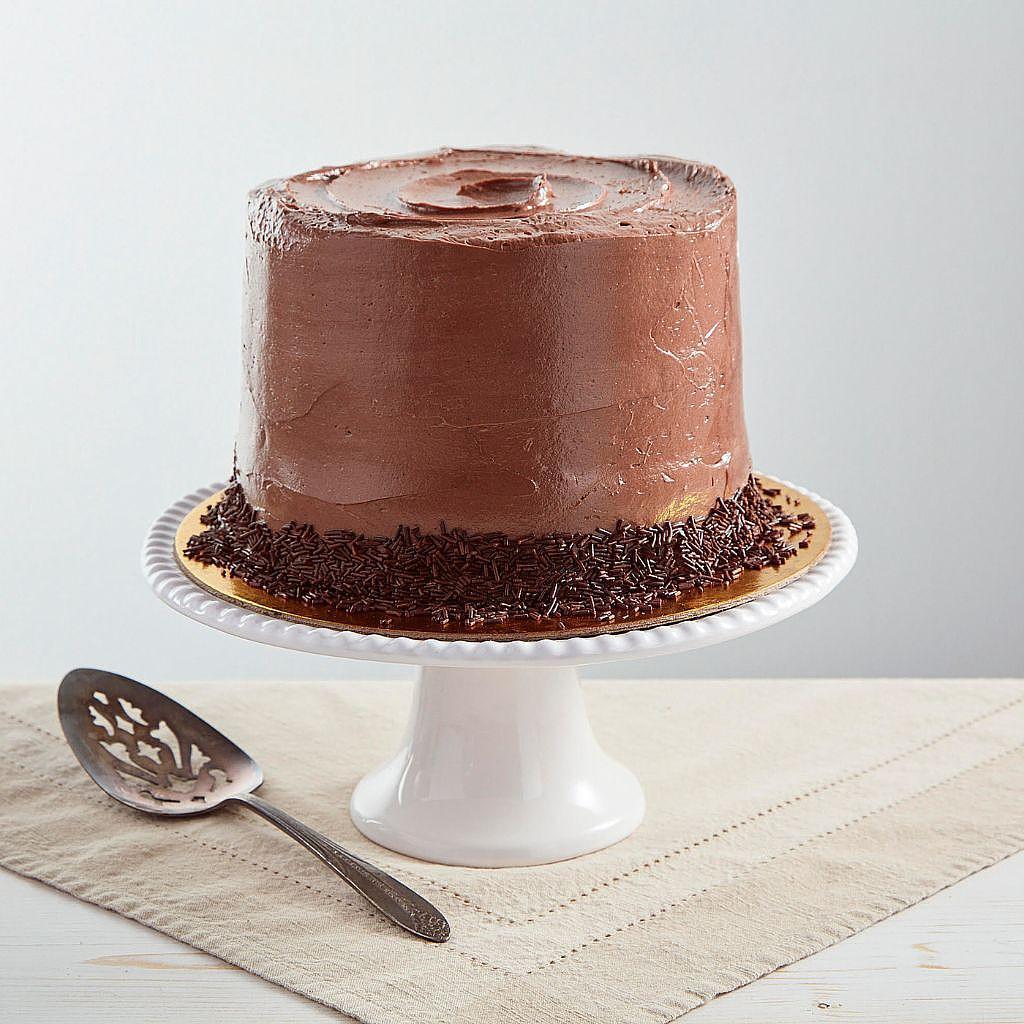 Six-inch vegan chocolate cake with smooth frosting and chocolate sprinkles on a white pedestal stand.